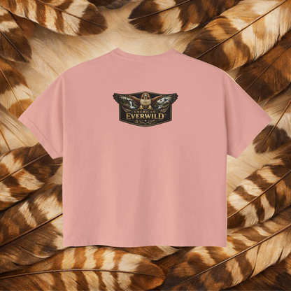 A peachy short-sleeve t-shirt with a central graphic of a hawk with wings outstreched with painted scene in the wings and the text 'American Everwild', displayed flat against a background of brown and tan feathers.