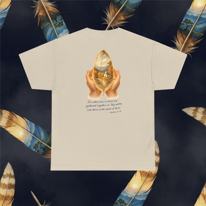 A sand color short-sleeve t-shirt displayed flat, showing a graphic on the back with a golden feather held by two hands and a Bible verse. The t-shirt is centered on a dark navy background decorated with large golden feathers.