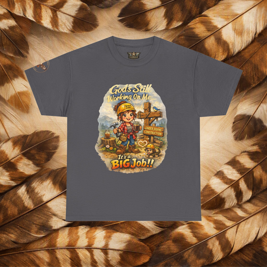 A dark gray crewneck t-shirt featuring a colorful cartoon graphic of a construction worker with a sign that reads 'Gods Still Working On Me It's a BIG Job!!', displayed flat against a background of brown and beige feathers.