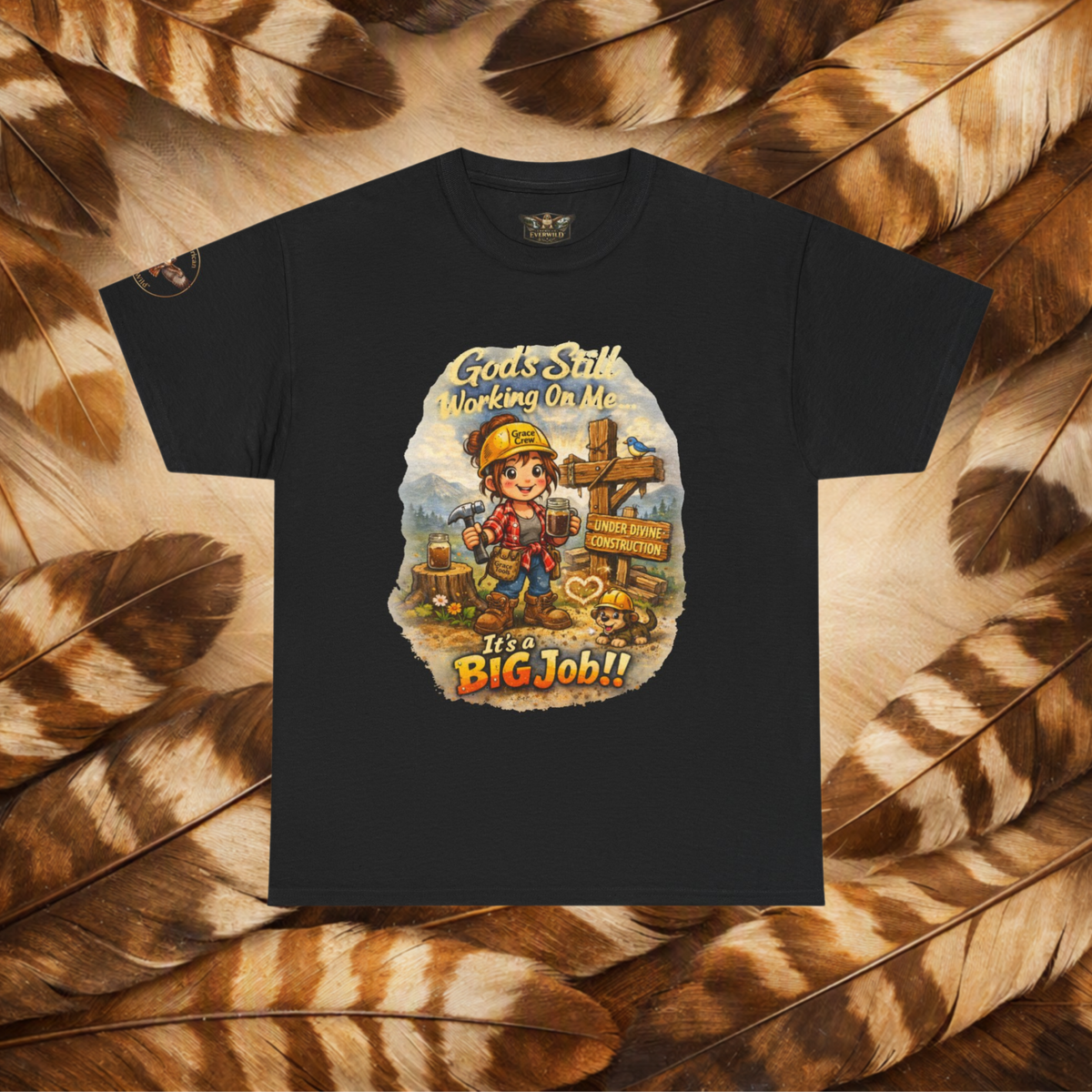 A black short-sleeve graphic t-shirt laid flat on a background of brown and beige feathers. The t-shirt features a colorful cartoon graphic of a construction worker with a hard hat and a sign that reads 'Gods Still Working On Me It's a BIG Job!!'.