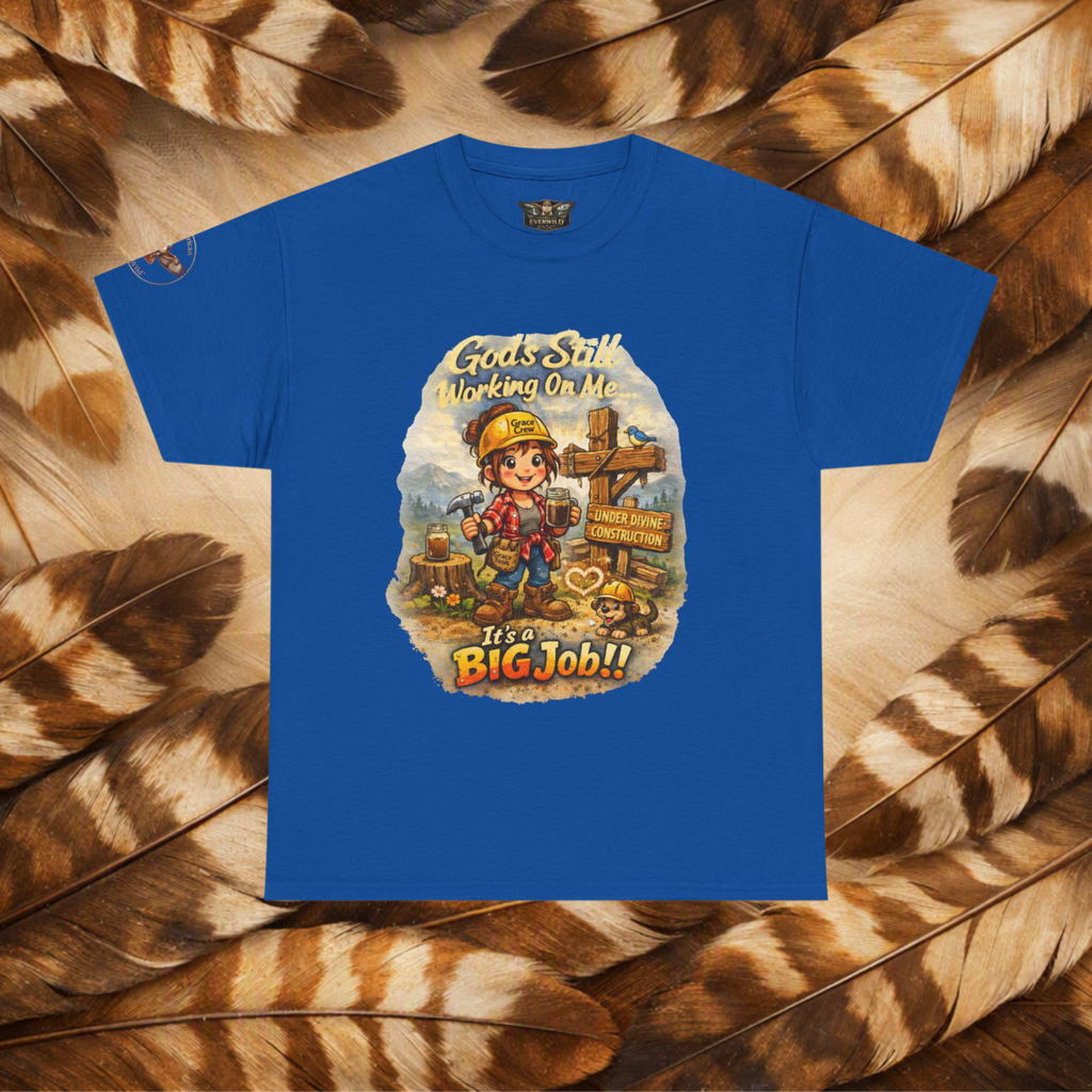A royal blue short-sleeve t-shirt with a colorful graphic print of a cartoon construction worker and the text 'Gods Still Working On Me It's a BIG Job!!', displayed flat against a background of brown and beige feathers.