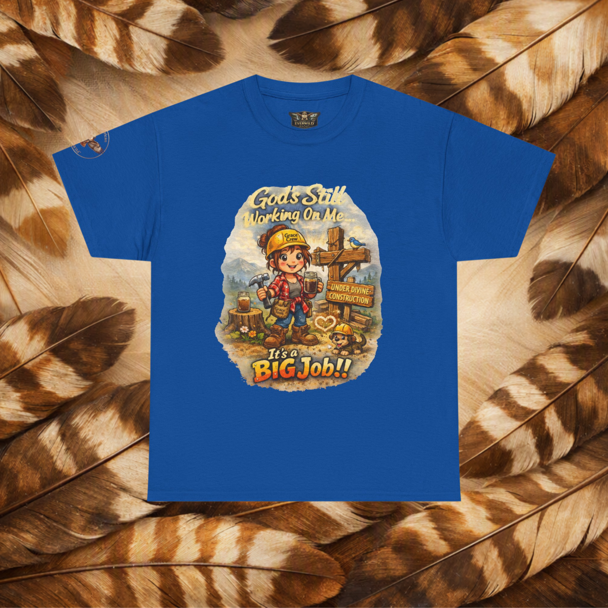 A royal blue short-sleeve t-shirt with a colorful graphic print of a cartoon construction worker and the text 'Gods Still Working On Me It's a BIG Job!!', displayed flat against a background of brown and beige feathers.