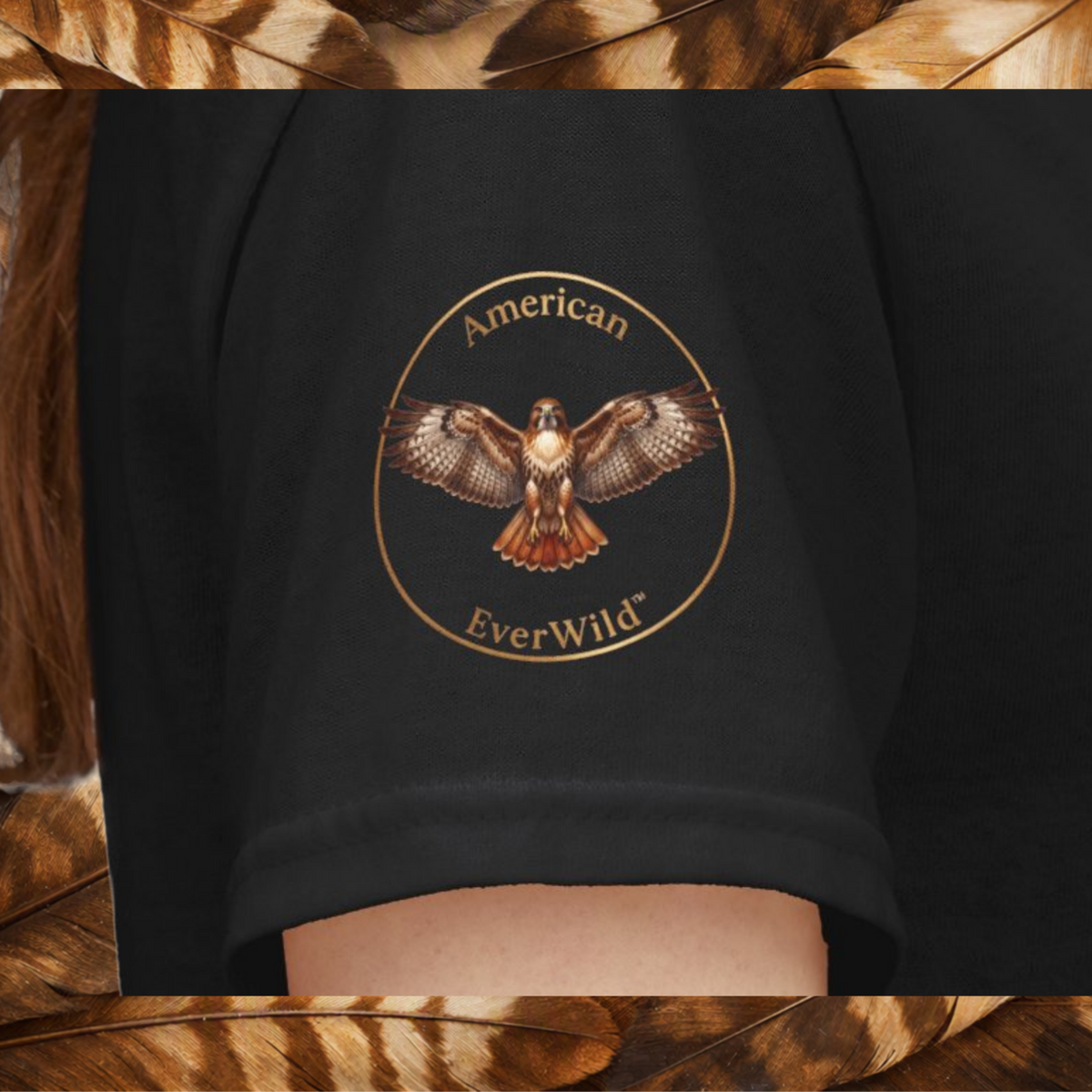 A close-up of a black sleeve featuring a circular graphic of a hawk with the text 'American EverWild' in gold, shown on a model on a back ground of brown and tan feathers.