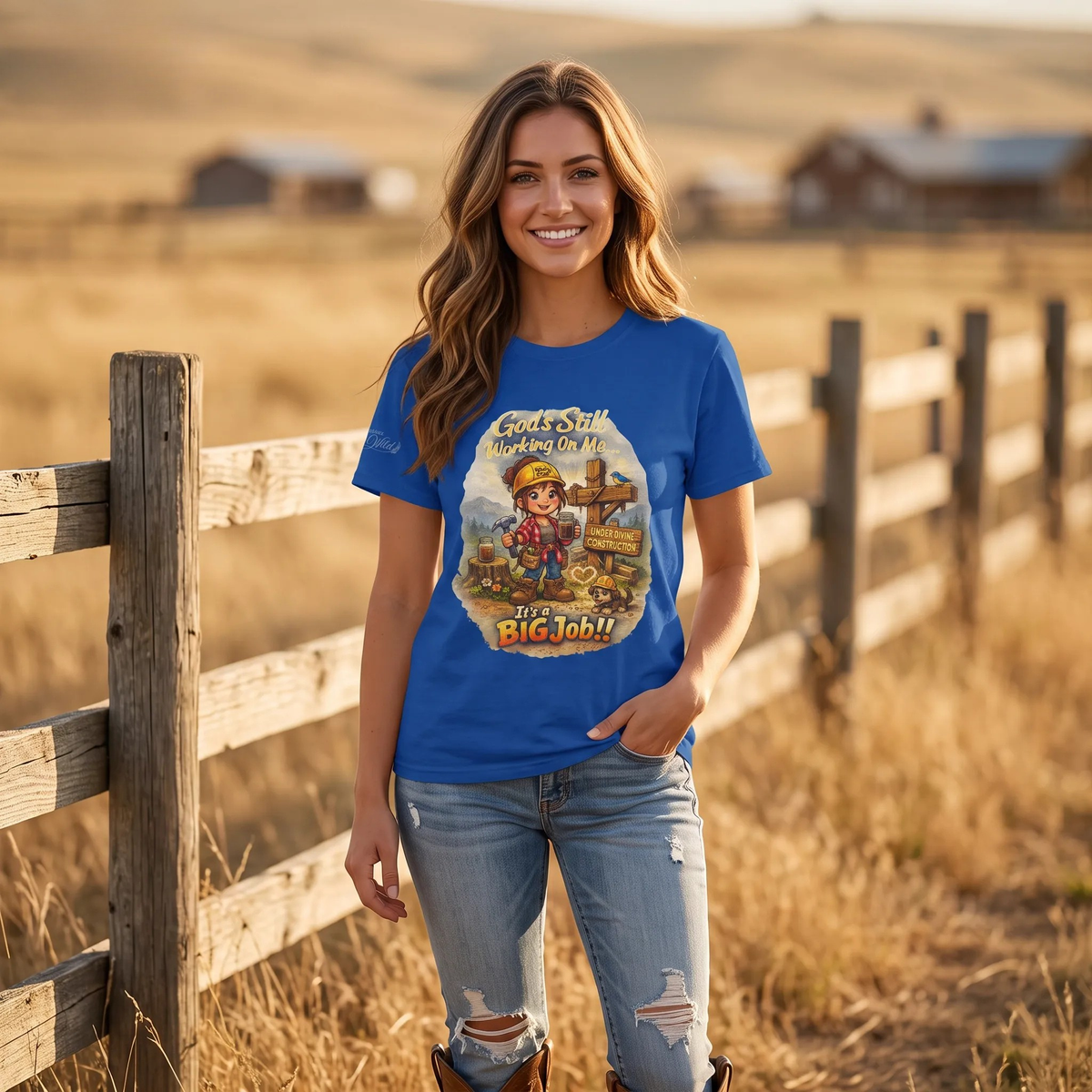 A woman stands outdoors in a dry grassy field beside a wooden fence, wearing a bright blue graphic T-shirt with a construction-themed illustration and the words 'God's Still Working on Me.' and 'It's A Big Job!' on the front of the t-shirt.