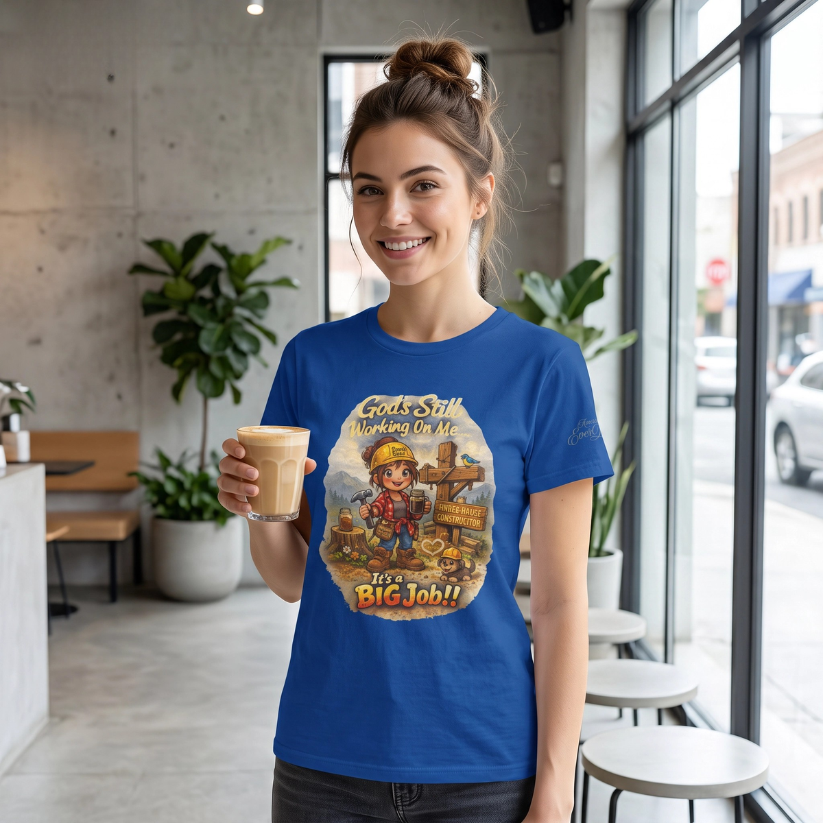 A woman stands in a bright modern cafe holding a latte and wearing a royal blue graphic t-shirt with a female cartoon construction character and the text 'Gods Still Working On Me—It's a Big Job!" on the front of the t-shirt.