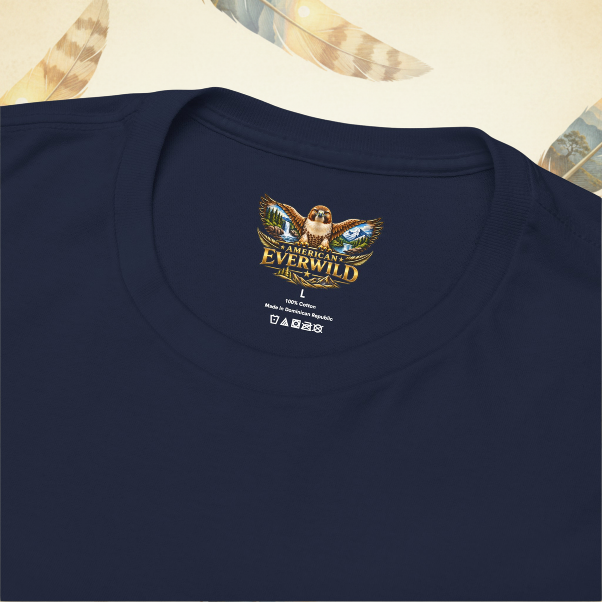 Close-up of a navy blue crewneck t-shirt showing the inner neck label with a colorful 'American Everwild' hawk graphic and care instructions, set against a light cream background with decorative feathers.