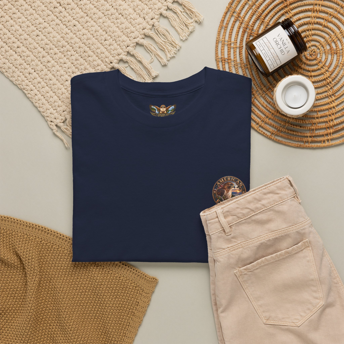 A navy blue folded t-shirt with printed graphic on the chest, styled in a flat lay with beige pants, a cream knit blanket, a brown candle jar, and a white mug on a light gray background.