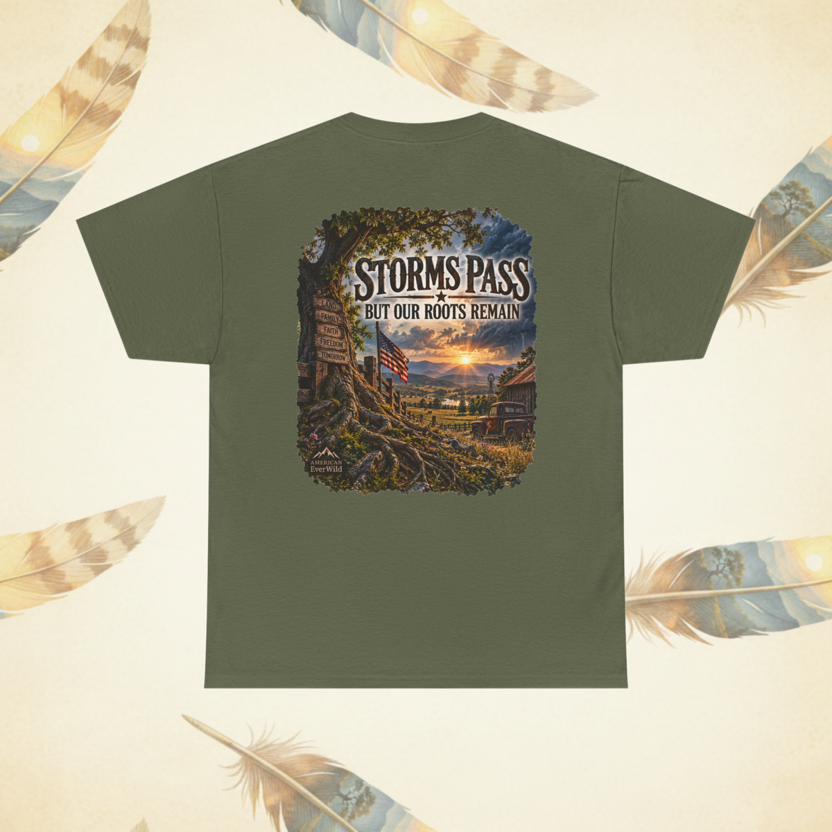 An olive green short-sleeve t-shirt shown from the back, featuring a large graphic print of a rustic mountain pass at sunset with the text 'STORMS PASS BUT OUR ROOTS REMAIN'. The shirt is displayed on a light cream background decorated with stylized feathers.