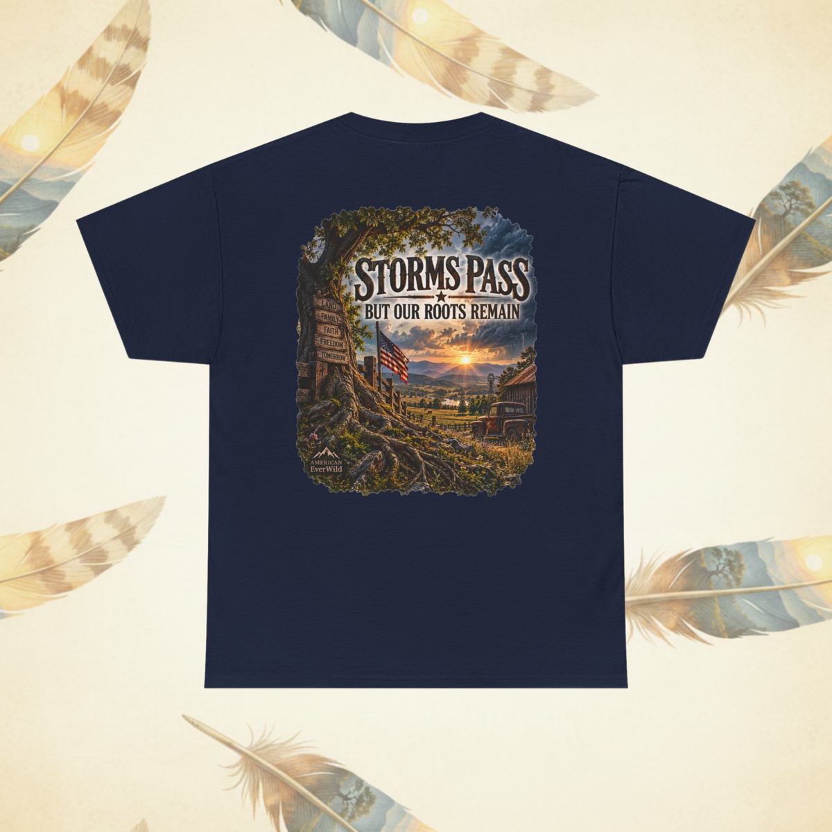 A navy blue short-sleeve t-shirt with a large rectangular graphic print on the back, featuring a scenic landscape illustration and the text 'STORMS PASS BUT OUR ROOTS REMAIN'. The t-shirt is displayed flat against a cream-colored background decorated with feather illustrations.