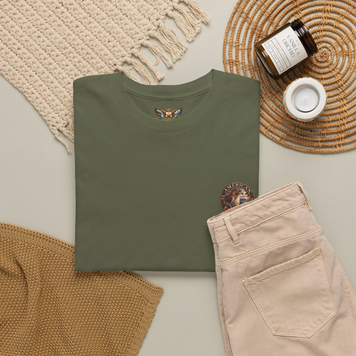 A miltary green folded t-shirt is displayed in a styled flat-lay arrangement alongside beige pants, a cream knit blanket, a brown textured throw, a small jar candle, and a white cup on a woven mat.