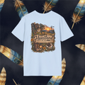 A light blue short-sleeve t-shirt with a detailed 'Heartland' western-themed graphic print on the front, featuring cowboy boots, a cowboy hat, a sunset, and a steer, displayed against a dark background decorated with feathers.
