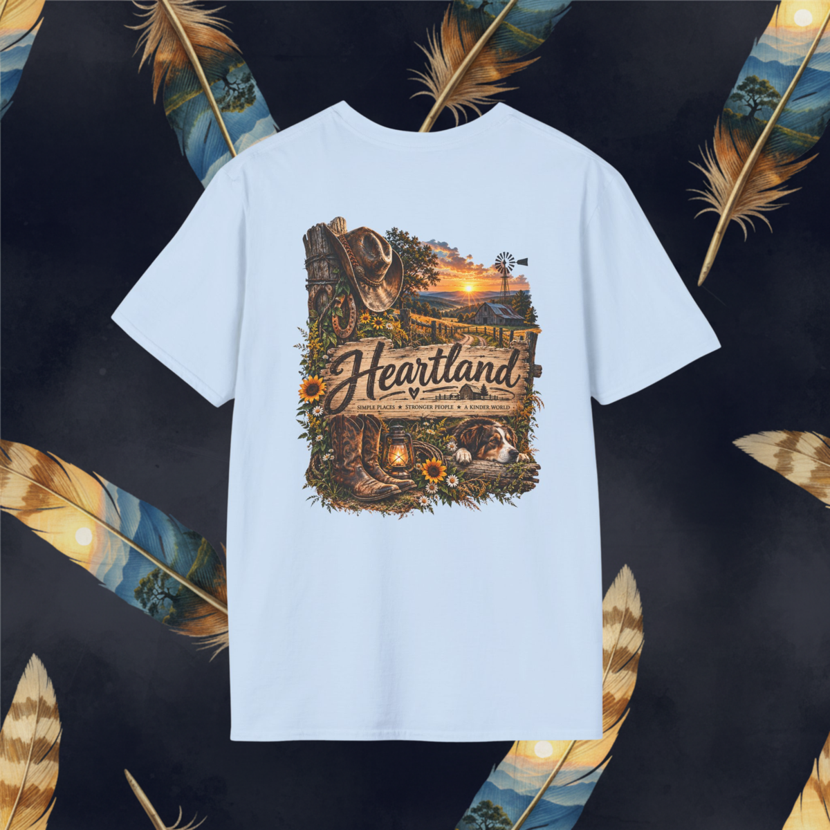 A light blue short-sleeve t-shirt with a detailed 'Heartland' western-themed graphic print on the front, featuring cowboy boots, a cowboy hat, a sunset, and a steer, displayed against a dark background decorated with feathers.