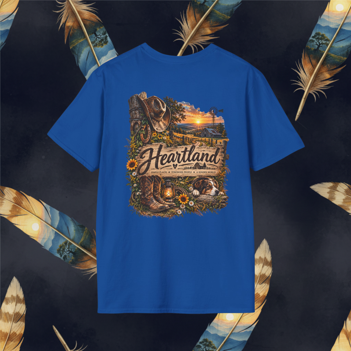 A royal blue short-sleeve t-shirt with a large, detailed graphic on the front depicting a rustic western farm scene with a cowboy hat, boots, lantern, and the text 'Heartland'. The shirt is displayed flat against a dark background decorated with large decorative feathers.