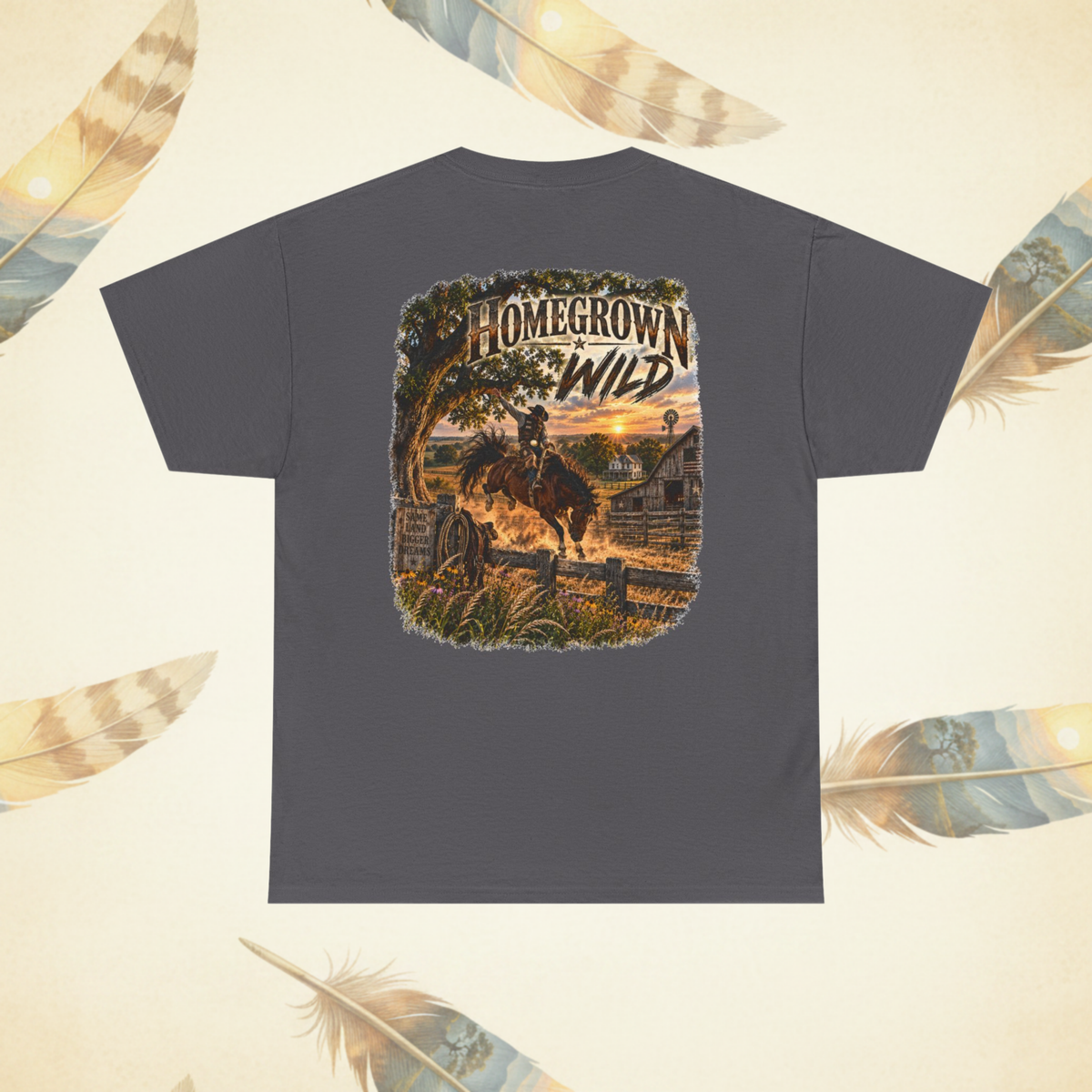 A dark gray t-shirt shown from the back, featuring a large graphic print of a cowboy riding a horse through a rustic western landscape at sunset with the text 'HOMEGROWN WILD'. The t-shirt is displayed against a light cream background decorated with scattered feather illustrations.