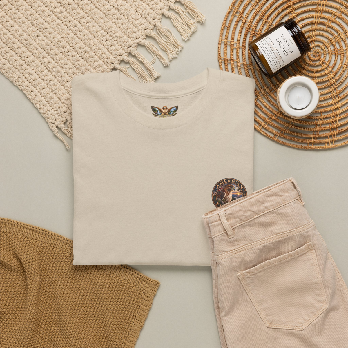 A folded Sand-colored t-shirt with a circular " American EverWild" logo  graphic on the upper left chest, styled in a flat lay with beige pants, a woven blanket, a candle, and a small cup on a neutral background.