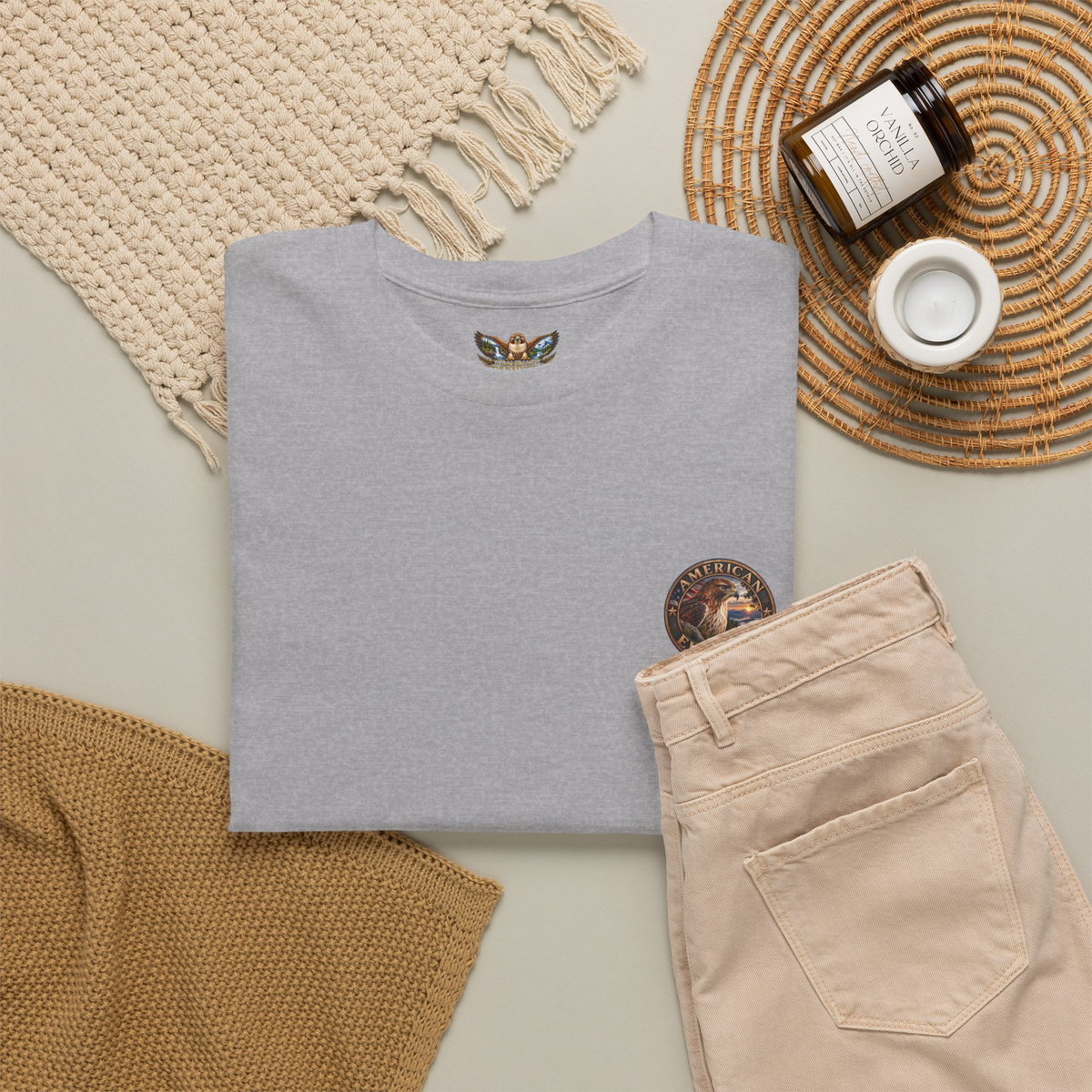 A folded light gray t-shirt with a small circular "American EverWild"
logo graphic on the upper left chest and a graphic hawk logo in the collar, styled in a flat lay with beige pants, a woven blanket, a candle, and a cup on a neutral background.
