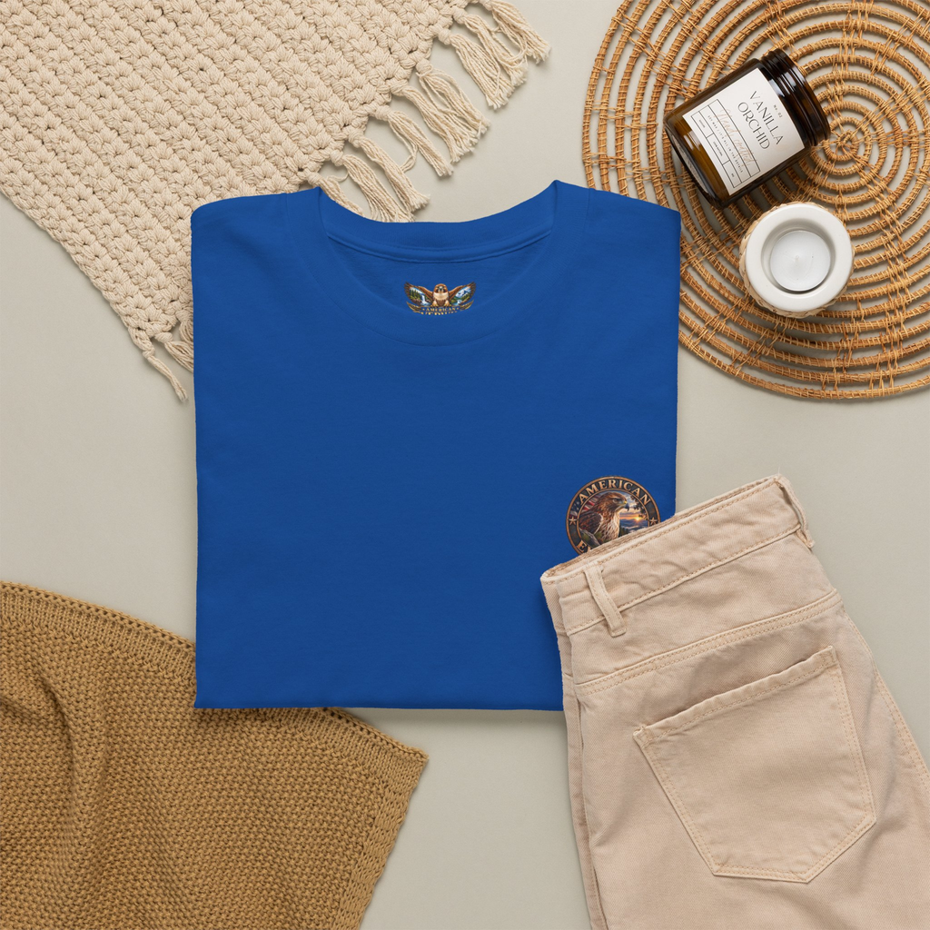 A folded royal blue t-shirt with a circular "American EverWild" logo graphic on the upper left chest, styled in a flat lay with beige pants, a cream knit blanket, a brown candle jar, and a white cup on a woven mat.