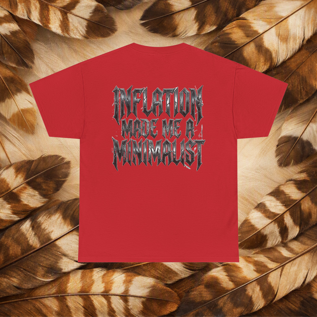A red short-sleeve t-shirt with a black and gray gothic-style graphic text reading 'INFLATION MADE ME A MINIMALIST', displayed flat on a background of brown and beige feathers.