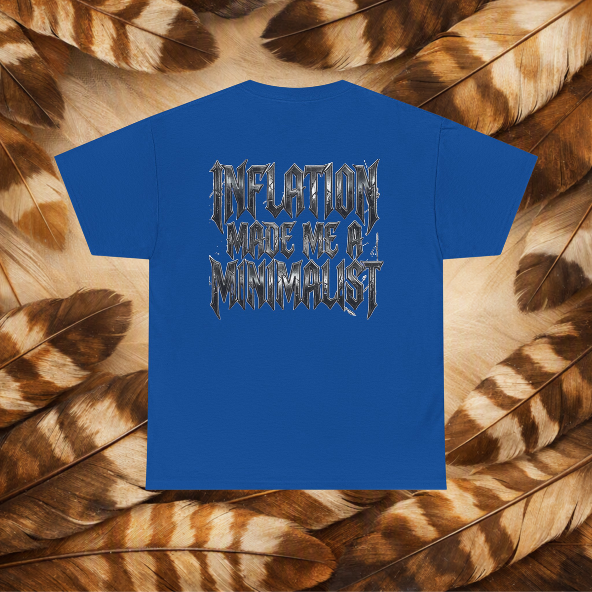A royal blue short-sleeve t-shirt with a stylized graphic on the back reading 'INFLATION MADE ME A MINIMALIST' in a metallic, jagged font. The shirt is laid flat on a background composed of brown and beige feathers.