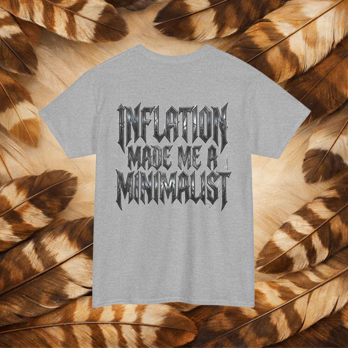 A sport gray t-shirt with a black gothic-style text graphic reading 'INFLATION MADE ME A MINIMALIST', displayed flat against a background of brown and beige feathers.