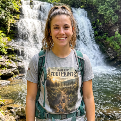 A smiling woman stands outdoors in front of a waterfall and pool, wearing a gray graphic T-shirt with the text 'LEAVE ONLY FOOTPRINTS' and a forest trail print, plus a green hiking backpack.