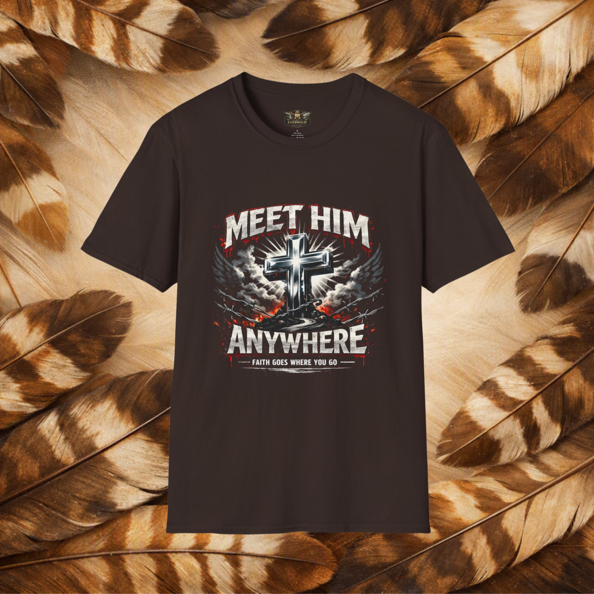 A dark brown graphic t-shirt displayed flat against a background of brown and beige feathers. The t-shirt features a graphic print with a metallic cross, wings, and the text 'MEET HIM ANYWHERE' in white and red.