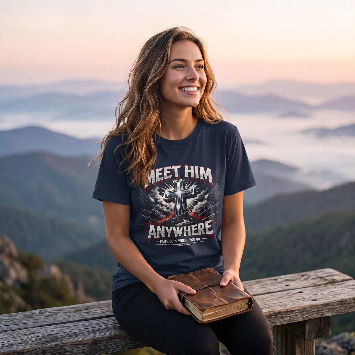 A woman sitting on a wooden railing outdoors at sunset, wearing a navy graphic T-shirt with the words 'MEET HIM ANYWHERE' and a cross design, holding a book.