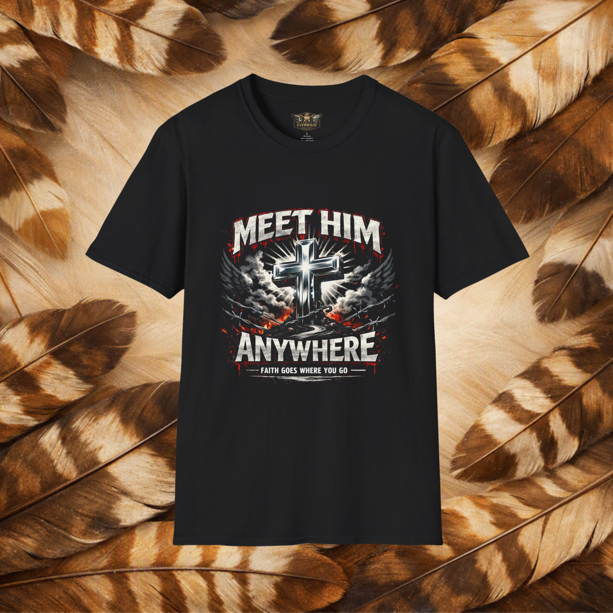 A black short-sleeve graphic t-shirt displayed flat against a background of brown and beige feathers. The shirt features a large front graphic with a metallic silver cross, white and red text reading 'MEET HIM ANYWHERE', and a banner that says 'FAITH GOES WHERE YOU GO'.