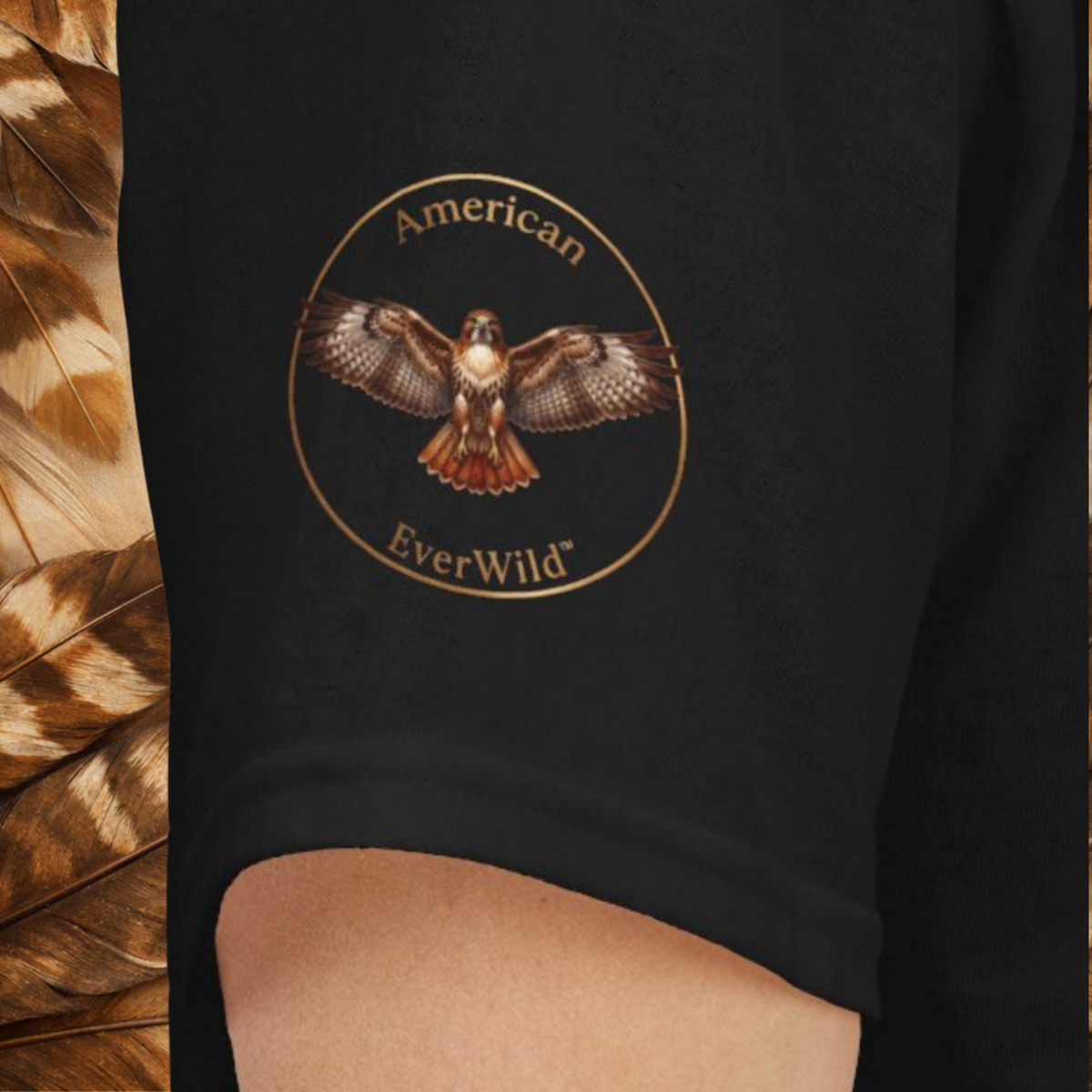 A close-up of a black garment featuring a circular 'American EverWild' logo with a hawk illustration in brown, gold, and white tones. A feather-patterned prop is visible on the left side.