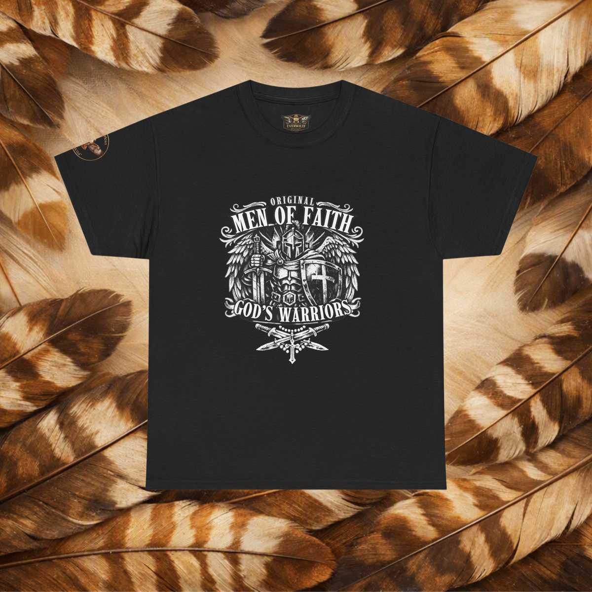 A black short-sleeve t-shirt with a white graphic print reading 'Original Men of Faith God's Warriors' and featuring a winged cross and dagger design, laid flat on a background of brown and tan feathers.