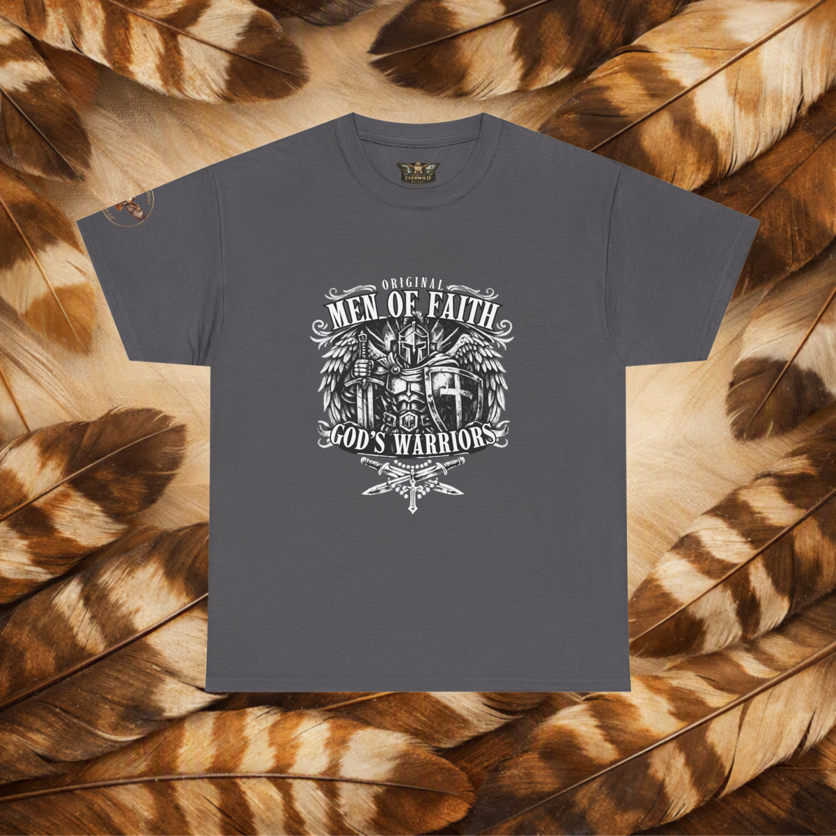A dark gray crewneck t-shirt with a white graphic print reading 'Original Men of Faith God's Warrior' surrounding a winged cross and eagle design, displayed flat on a background of brown and tan feathers.