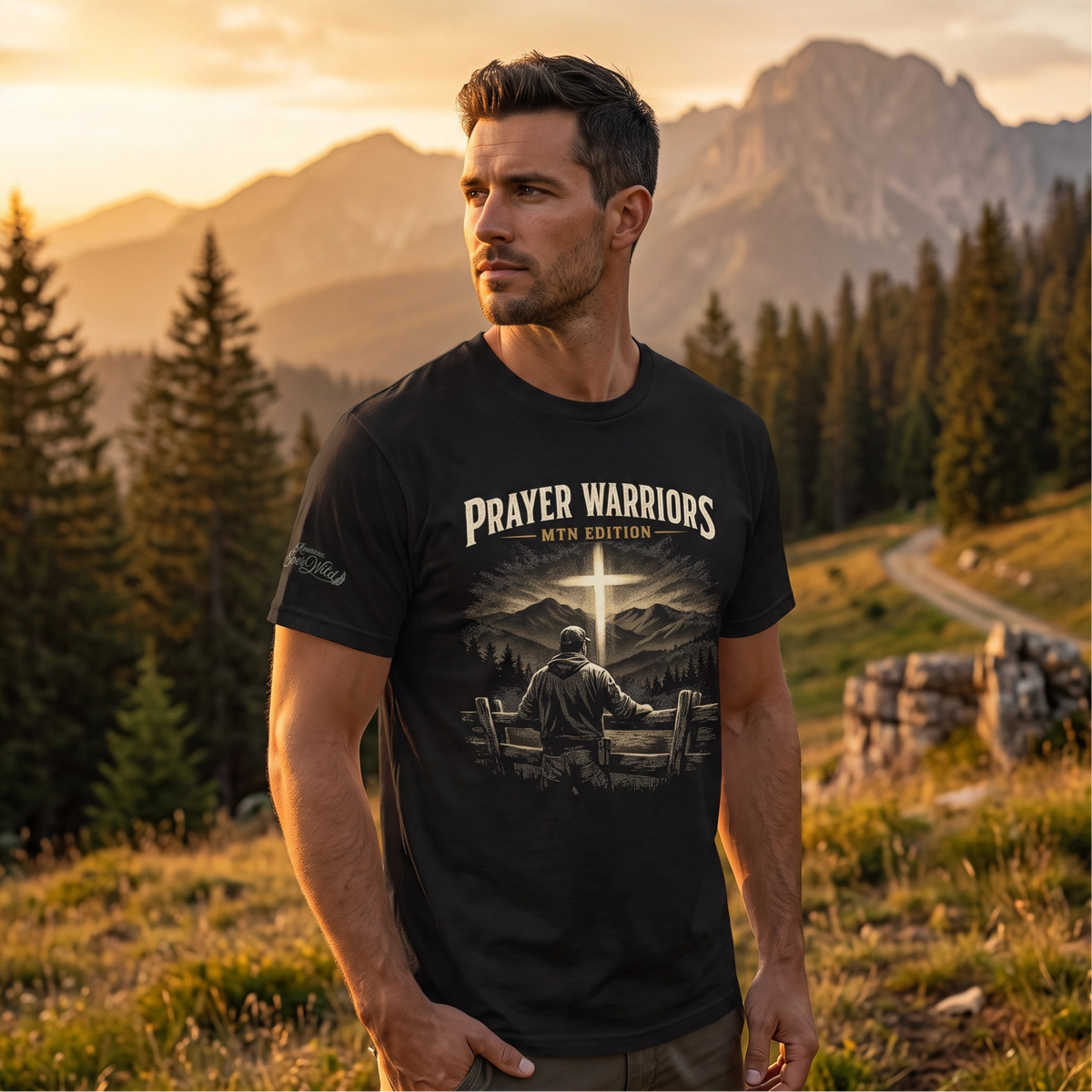 A man wearing a black graphic t-shirt with 'PRAYER WARRIORS MTN EDITION' printed on the front, standing in a scenic mountain and pine forest during golden hour.