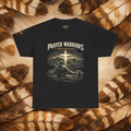A black graphic t-shirt featuring the text 'PRAYER WARRIORS - MTN EDITION' above an illustration of a person leaning on a fence with a cross and mountains in the background. The shirt is displayed flat against a background of brown and beige feathers.