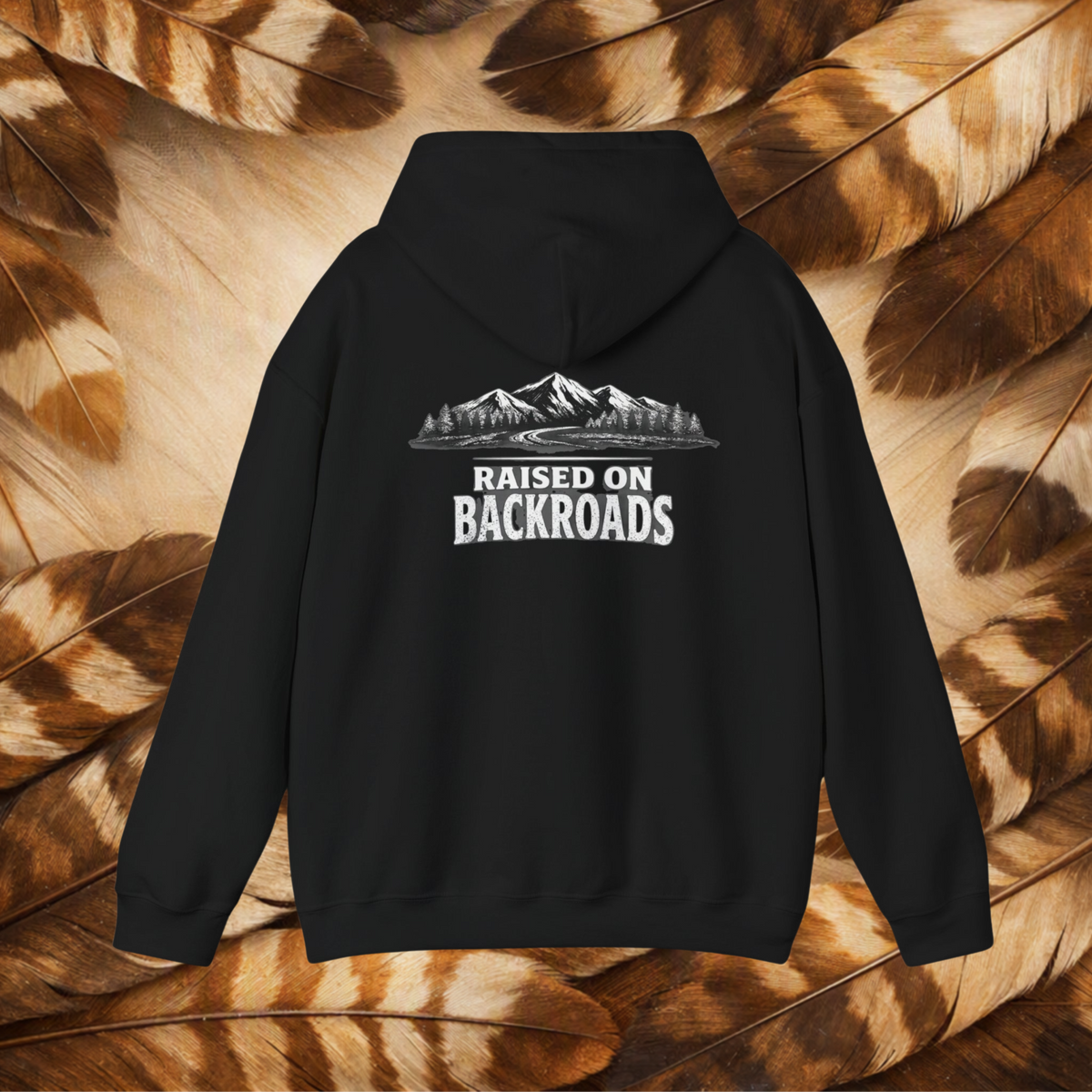 A black pullover hoodie shown from the back, featuring a white graphic print of mountains, trees, and the text 'RAISED ON BACKROADS' centered on the back. The hoodie is displayed flat against a background composed of brown and tan feathers.