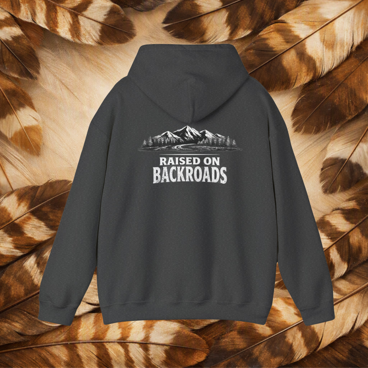 A charcoal hoodie shown from the back, featuring a white graphic of mountains, trees, and a road with the text 'RAISED ON BACKROADS'. The hoodie is displayed flat against a background of brown and beige feathers.