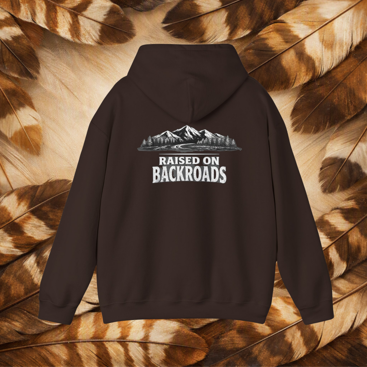 A chocolate brown pullover hoodie shown from the back, featuring a white graphic print of mountains, trees, and the text 'RAISED ON BACKROADS'. The hoodie is displayed flat against a background composed of brown and beige feathers.