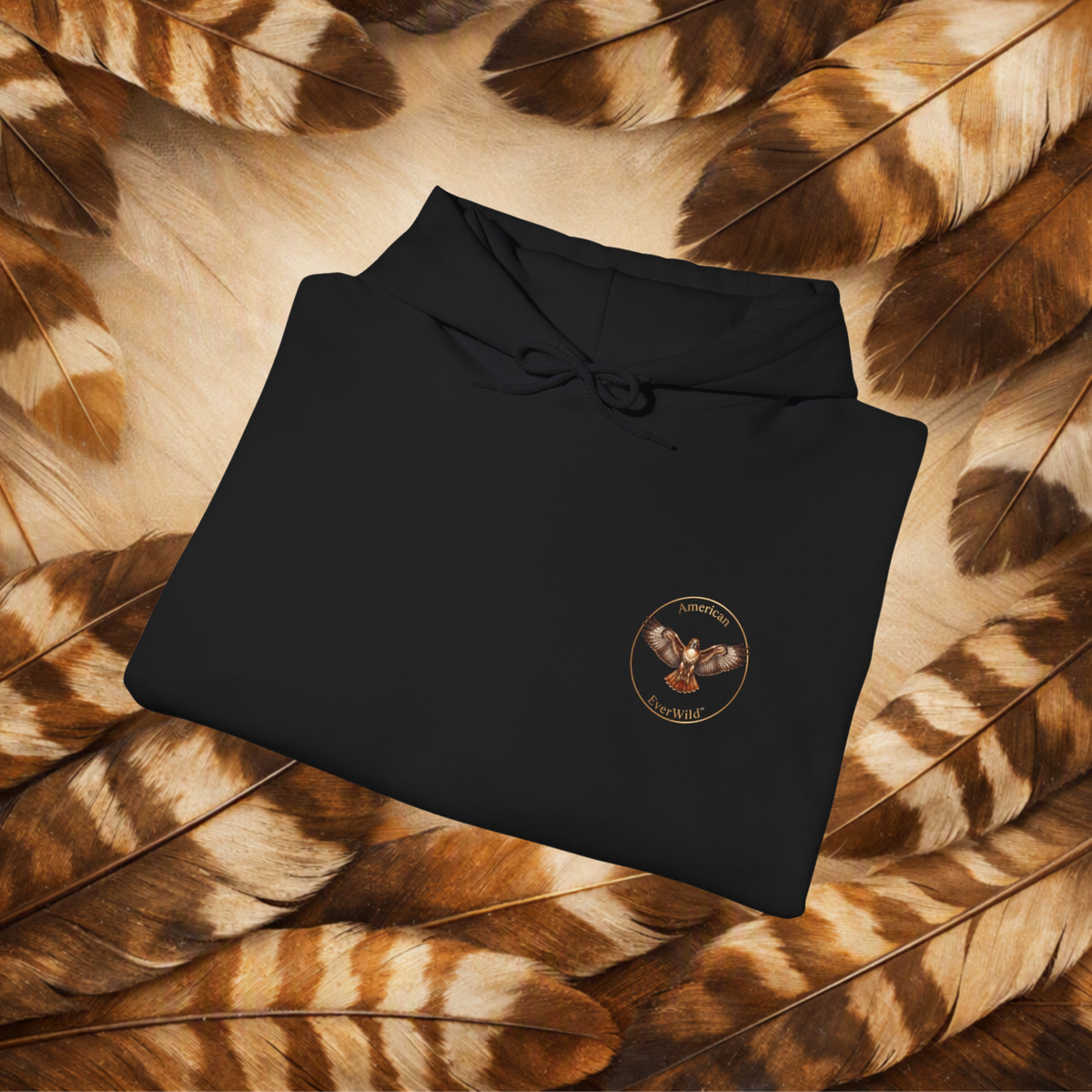 A folded black hoodie with a drawstring hood and a circular chest graphic for American EverWild, placed on a bed of brown and beige feathers. The graphic shows a red-tailed hawk