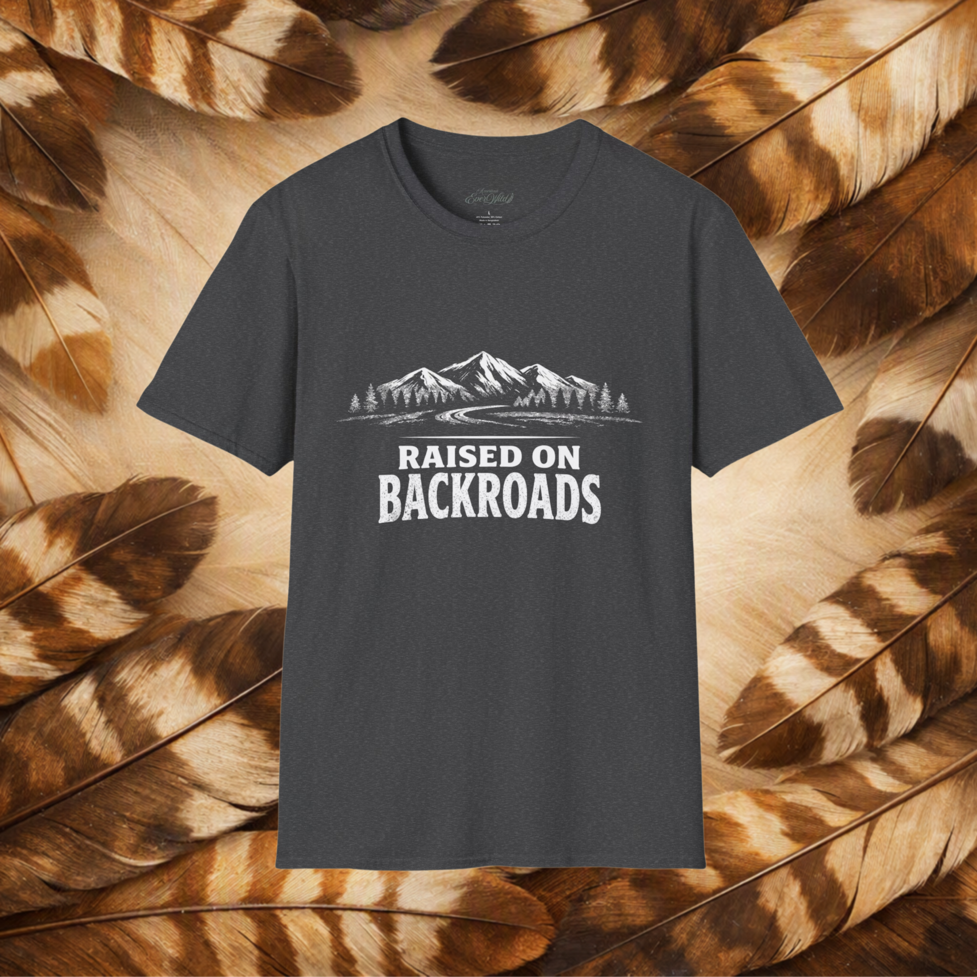 A dark charcoal gray t-shirt with a white graphic of mountains, trees, and the text 'RAISED ON BACKROADS' printed on the chest. The t-shirt is displayed flat against a background composed of brown and beige feathers.