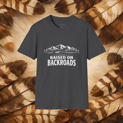 A dark charcoal gray t-shirt with a white graphic of mountains, trees, and the text 'RAISED ON BACKROADS' printed on the chest. The t-shirt is displayed flat against a background composed of brown and beige feathers.