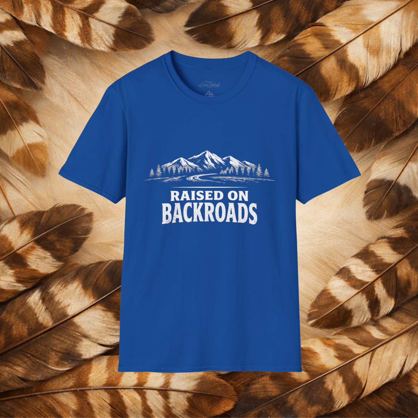 A royal blue short-sleeve t-shirt with a white graphic of mountains, trees, and the text 'RAISED ON BACKROADS' on the chest, displayed flat against a background of brown and beige feathers.