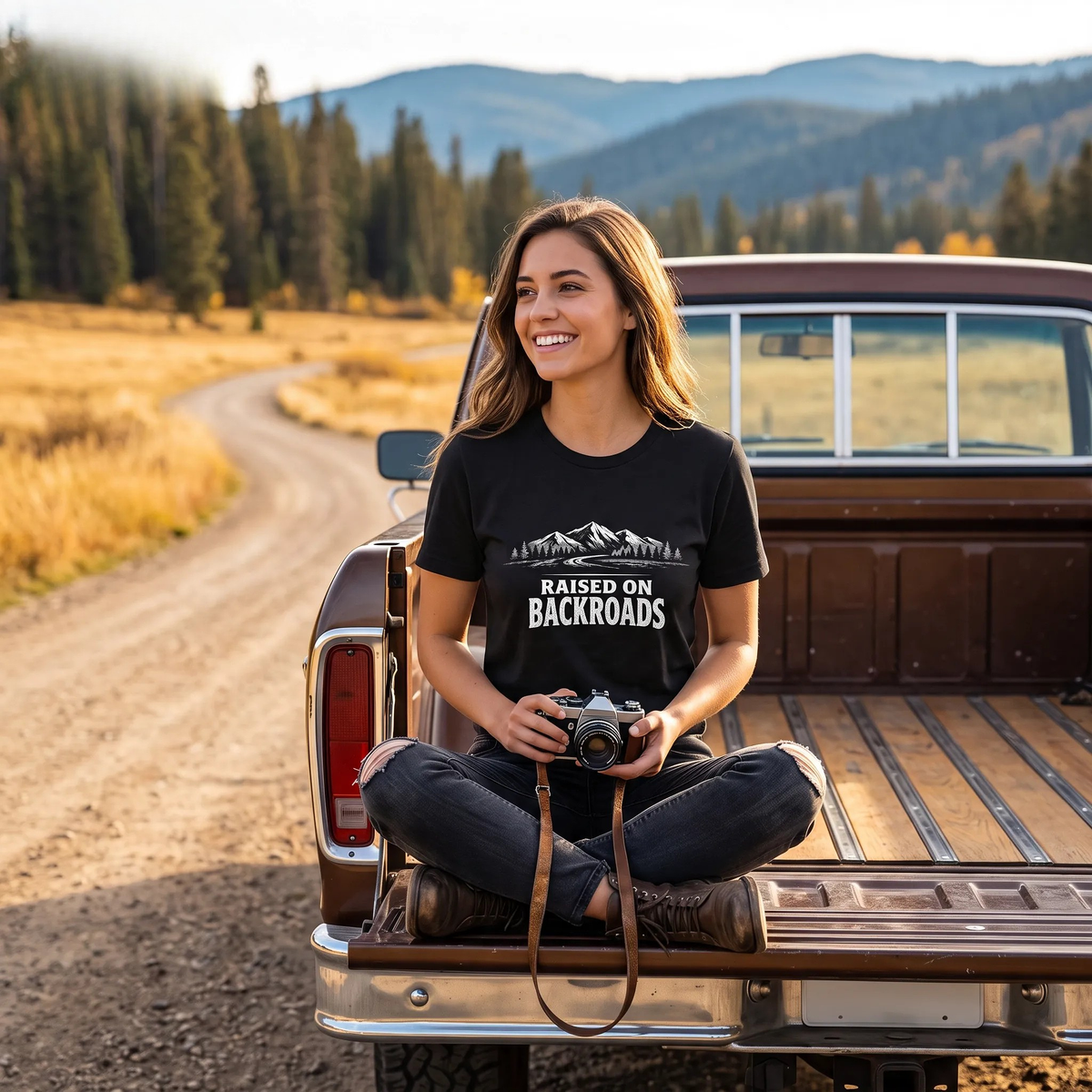 A woman sits cross-legged in the bed of a vintage pickup truck on a dirt road, wearing a black graphic t-shirt that reads 'RAISED ON BACKROADS' with a mountain design. The background shows a scenic mountain landscape with pine trees and golden fields.