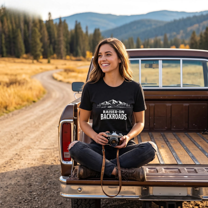 A woman sits cross-legged in the bed of a vintage pickup truck on a dirt road, wearing a black graphic t-shirt that reads 'RAISED ON BACKROADS' with a mountain design. The background shows a scenic mountain landscape with pine trees and golden fields.