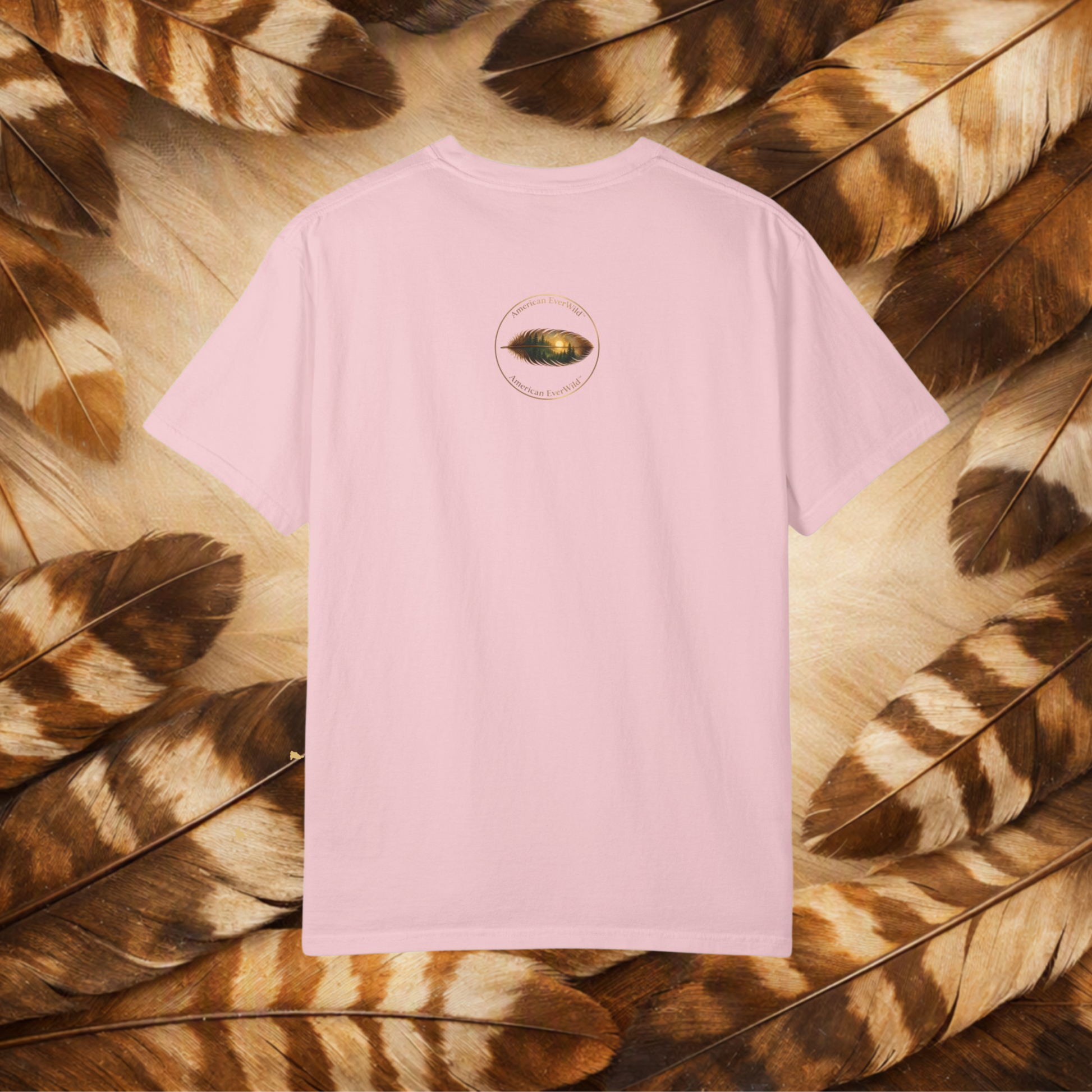 A light pink short-sleeve t-shirt with a small circular graphic of a painted feather with "American EverWild" on the center back, laid flat on a background of brown and beige feathers.