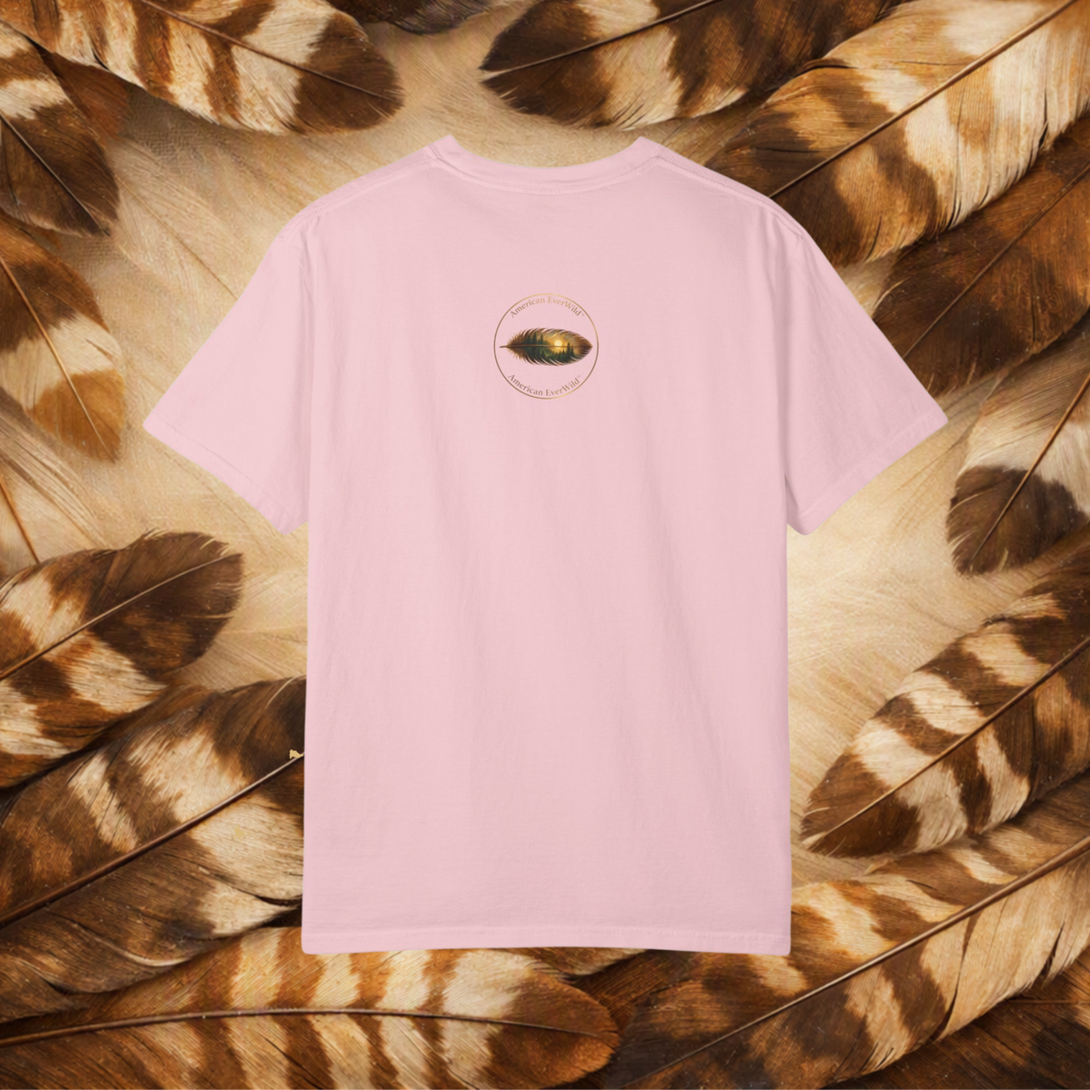 A light pink short-sleeve t-shirt with a small circular graphic of a painted feather with "American EverWild" on the center back, laid flat on a background of brown and beige feathers.