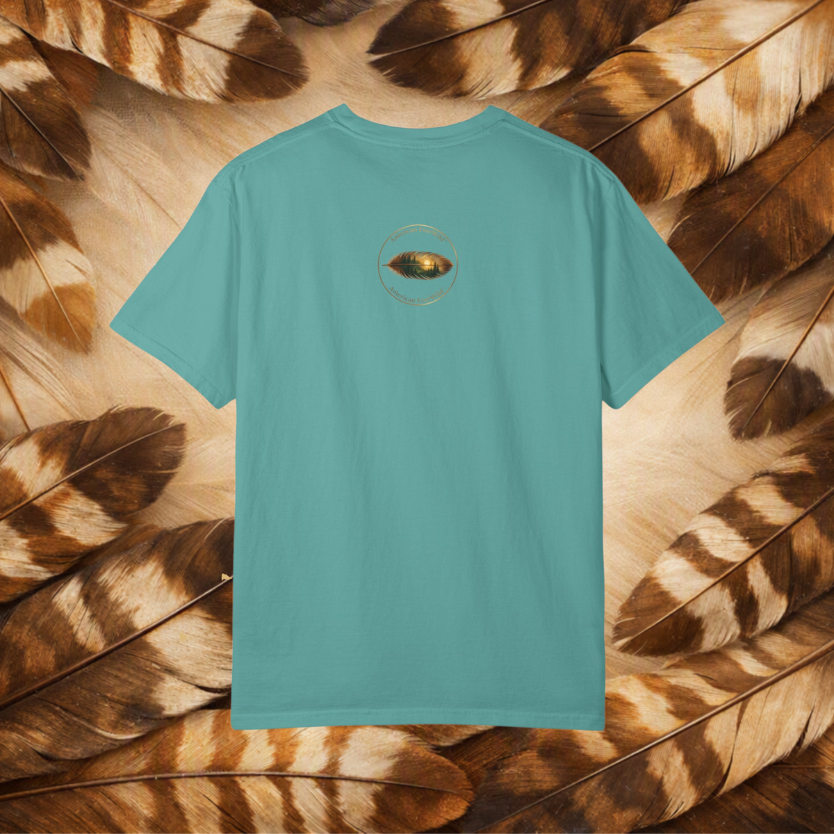A turquoise short-sleeve t-shirt shown from the back, featuring a small circular graphic of a painted feather in the center chest. The shirt is laid flat on a background composed of brown and beige feathers.