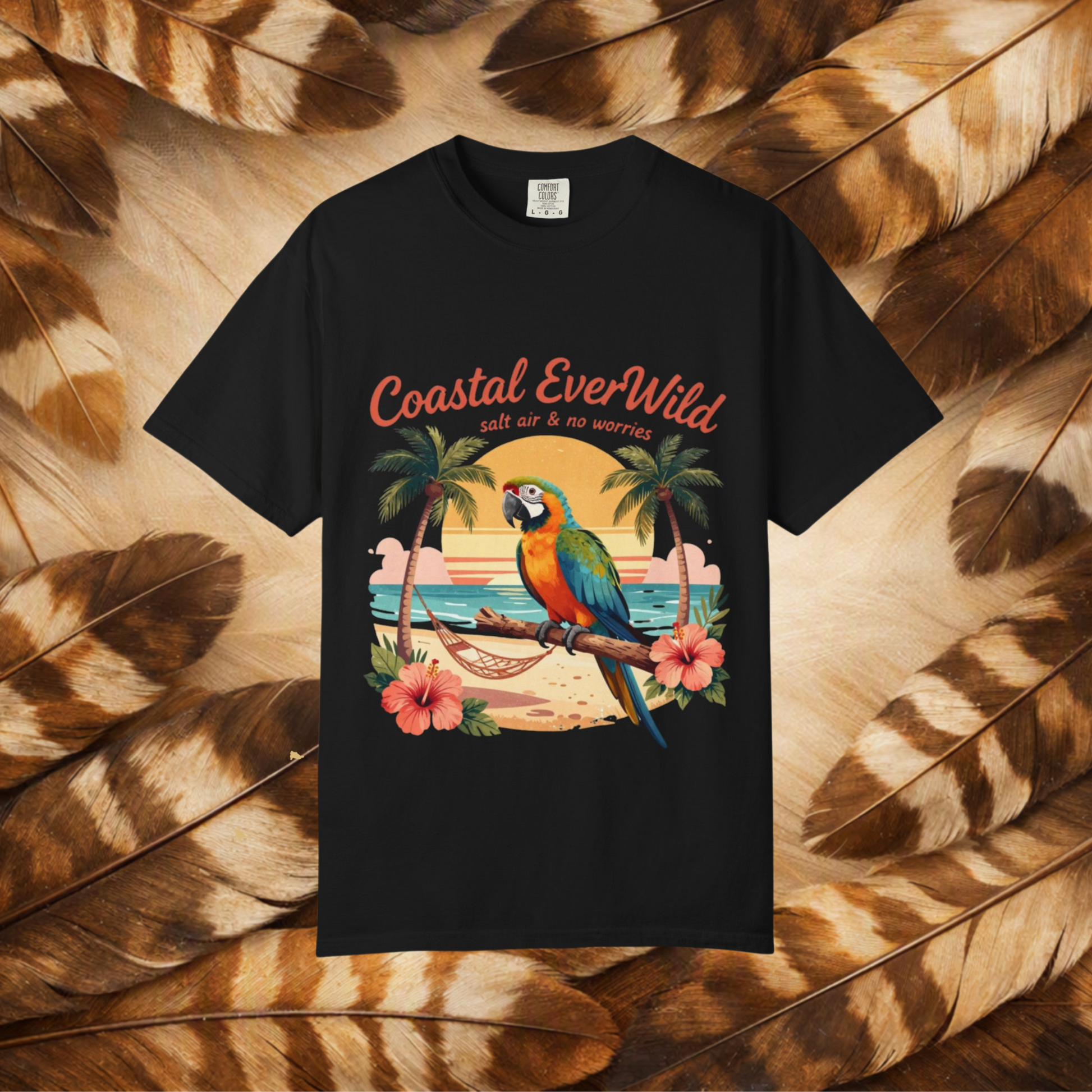 A black short-sleeve t-shirt with a colorful graphic print of a parrot on a beach and the text 'Coastal EverWild' and 'salt air & no worries', displayed against a background of brown and beige feathers.