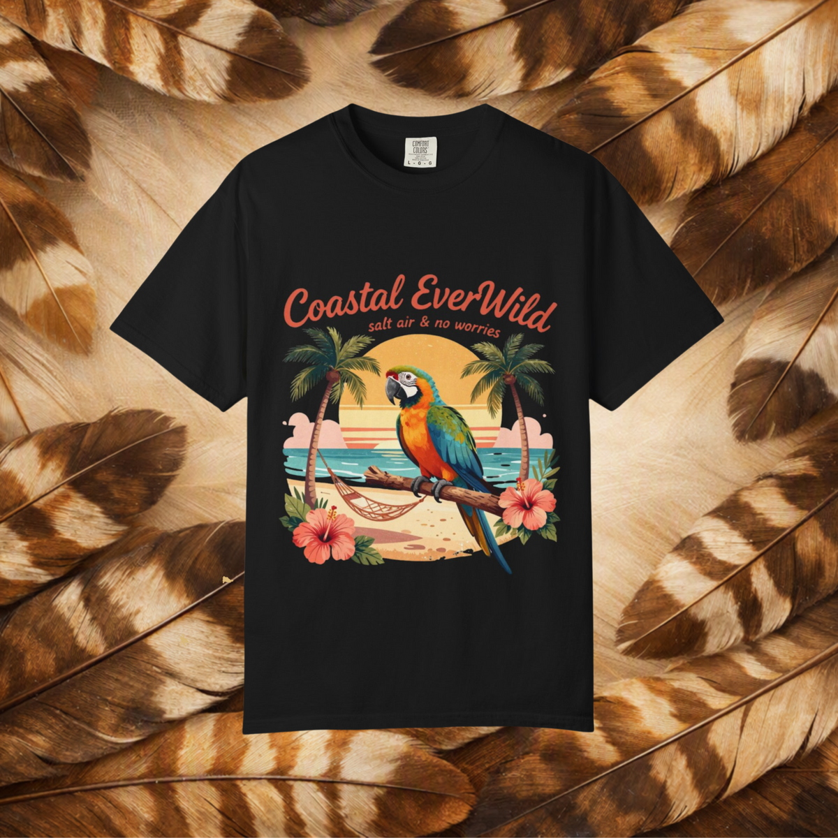 A black short-sleeve t-shirt with a colorful graphic print of a parrot on a beach and the text 'Coastal EverWild' and 'salt air & no worries', displayed against a background of brown and beige feathers.