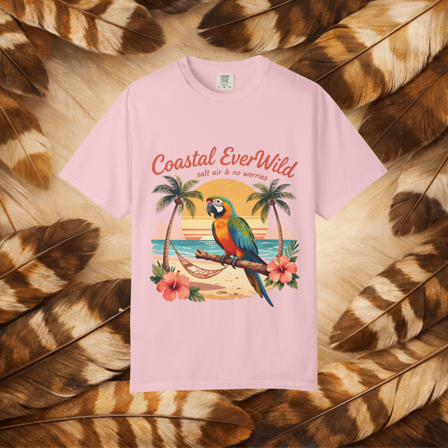 A light pink short-sleeve t-shirt with a colorful graphic of a parrot on a beach, surrounded by palm trees and hibiscus flowers, with the text 'Coastal EverWild salt air & no worries'. The shirt is laid flat on a background of brown and beige feathers.