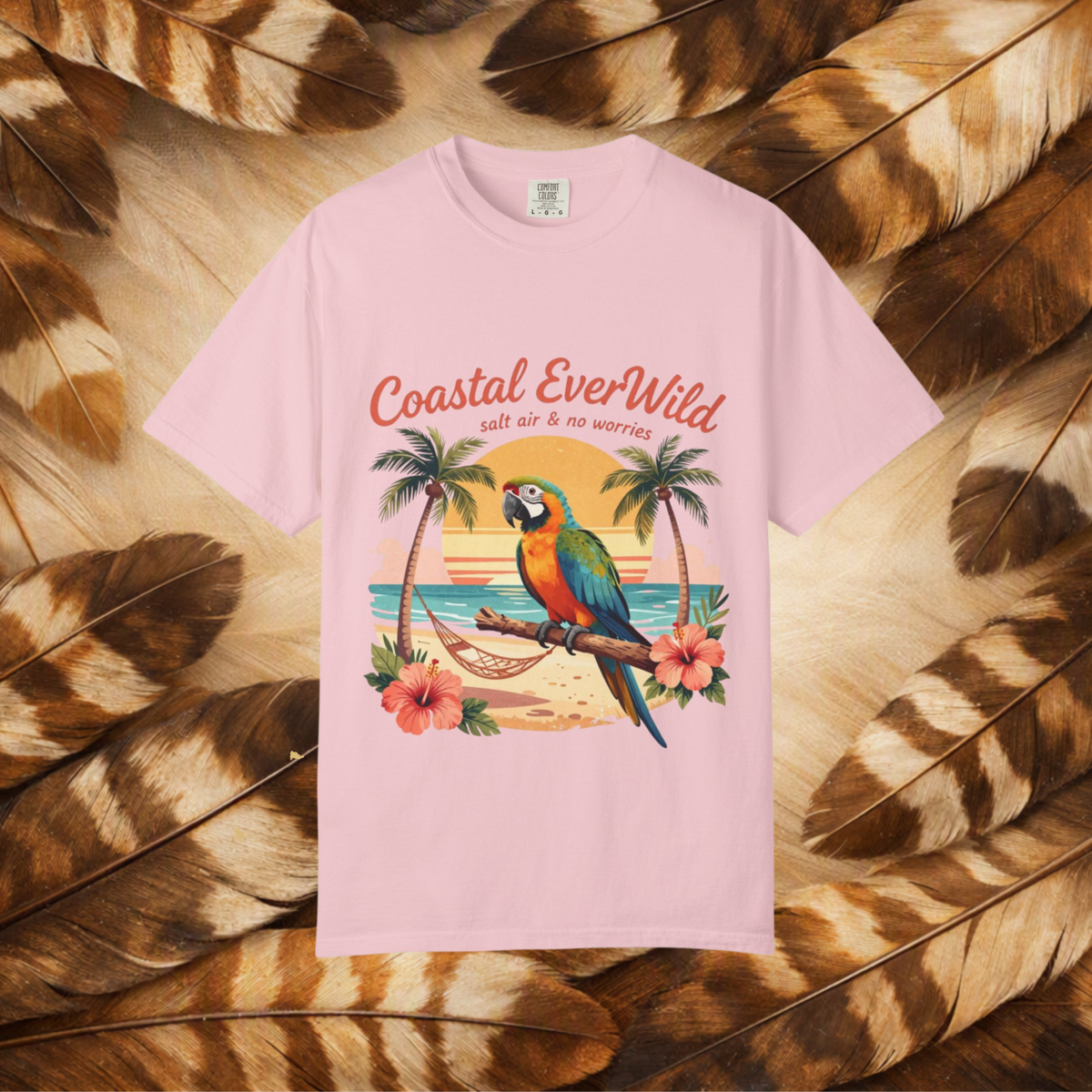 A light pink short-sleeve t-shirt with a colorful graphic of a parrot on a beach, surrounded by palm trees and hibiscus flowers, with the text 'Coastal EverWild salt air & no worries'. The shirt is laid flat on a background of brown and beige feathers.
