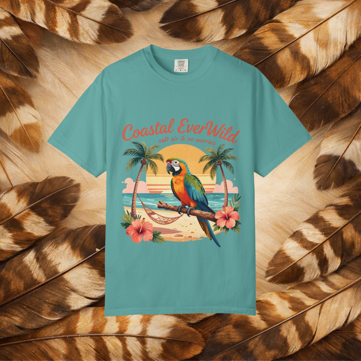 A turquoise short-sleeve t-shirt with a colorful graphic of a parrot on a beach and the text 'Coastal EverWild', presented flat against a background of brown and tan feathers.